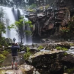 A wide shot of Phantom Falls cascading down dark, mossy cliffs into a rocky river, with several tourists visible, some taking photos and others observing the scene.