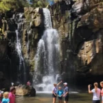 A bright, sunny scene of Phantom Falls flowing into a calm pool, with several families of tourists enjoying the water and surrounding rocks, some posing for photos.