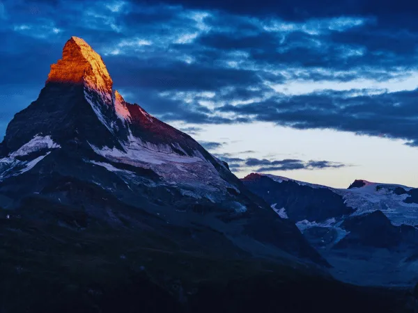 Matterhorn mountain peak glowing orange and gold at sunrise against dark dramatic clouds over the Swiss Alps near Zermatt