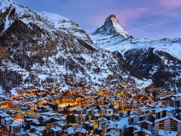 Aerial view of Zermatt village lit up at dusk in winter with the Matterhorn towering behind snow-covered rooftops