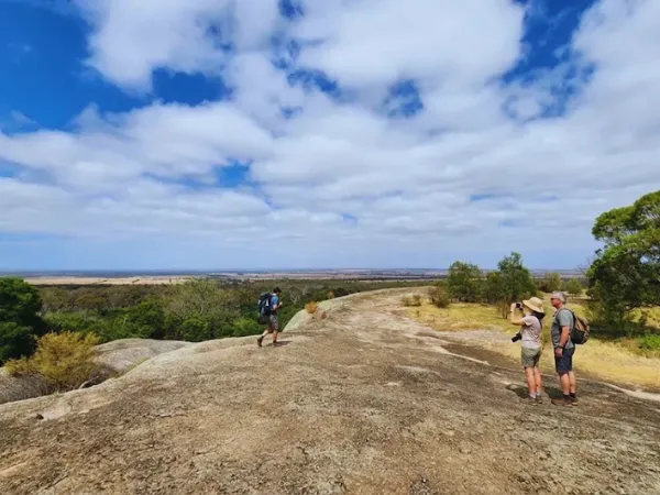 Three hikers standing on the granite summit of Big Rock at You Yangs Regional Park with wide plains and blue sky stretching to the horizon.