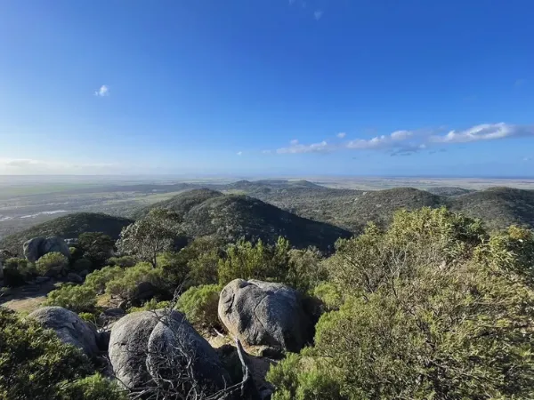 Panoramic view from You Yangs Regional Park summit showing rolling hills covered in green bushland with flat plains and coastline in the distance under a blue sky.
