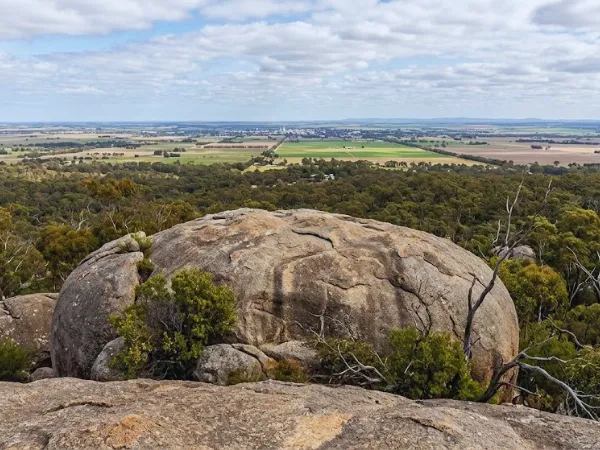 Large rounded granite boulder at Big Rock You Yangs Regional Park with expansive views of green farmland, eucalyptus woodland, and a distant township under a partly cloudy sky.
