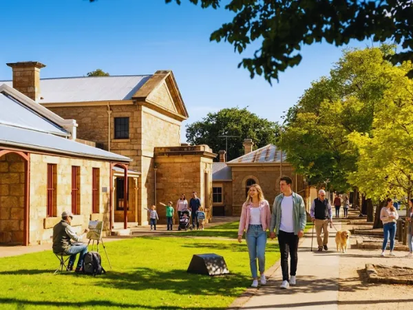 Couple and visitors walking through the tree-lined grounds of Beechworth's historic sandstone precinct with colonial buildings and a painter in the foreground on a sunny day