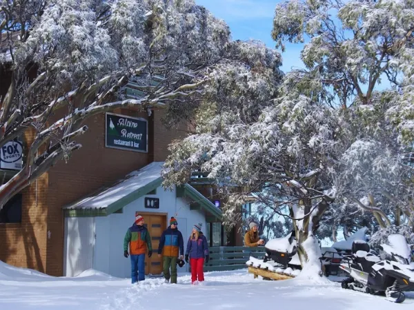 Three visitors standing outside the Alpine Retreat restaurant and bar at Mt Buller Village surrounded by deep snow and snow-laden gum trees in winter
