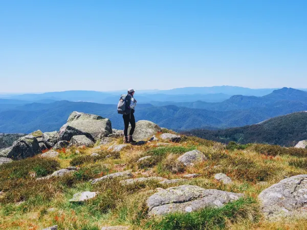 A hiker with a backpack standing on rocky alpine terrain at the summit of the Summit Nature Walk Mt Buller with vast mountain ranges stretching into the distance