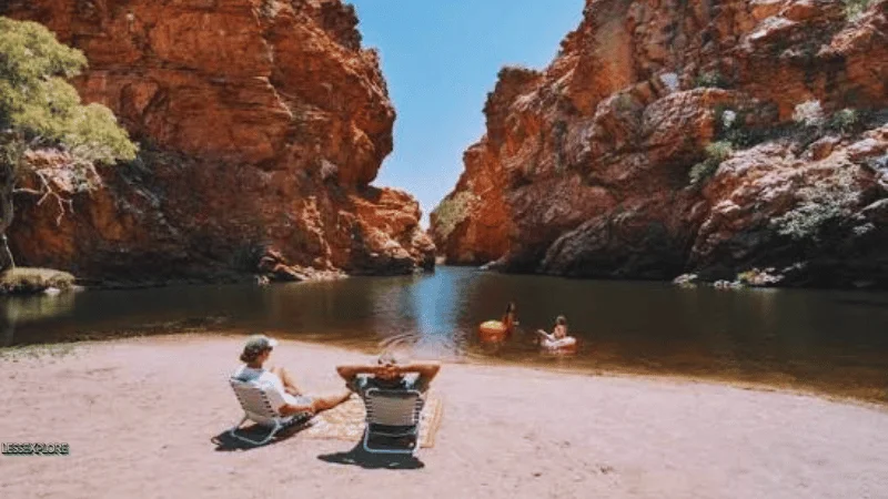 Visitors relaxing in camp chairs on the sand while two people float on inflatables at Ormiston Gorge